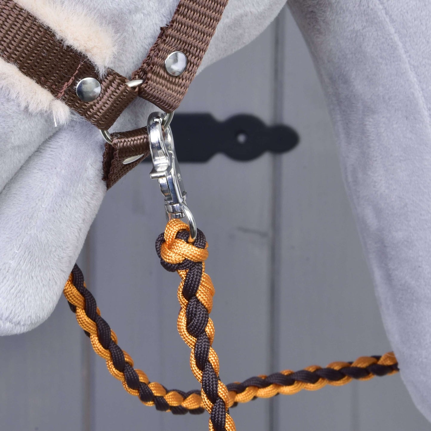 Detailní záběr na koňskou hlavu se Set Perfect Combination Brown od Hobby Horse LarDen, s ohlávkou s kožešinovým polstrováním a oranžovo-hnědým pleteným vodítkem se stříbrnou karabinou. V pozadí je rozmazané šedé dveře s černým štítkem klíčové dírky – ideální pro fanoušky hobby horse.