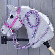 Set Perfect Combination Pink od Hobby Horse LarDen, šedý plyšový Hobby Horse s růžovou a tyrkysovou ohlávkou s kožešinovým polstrováním a otěžemi, visí na nástěnném háčku před šedým dřevěným pozadím.
