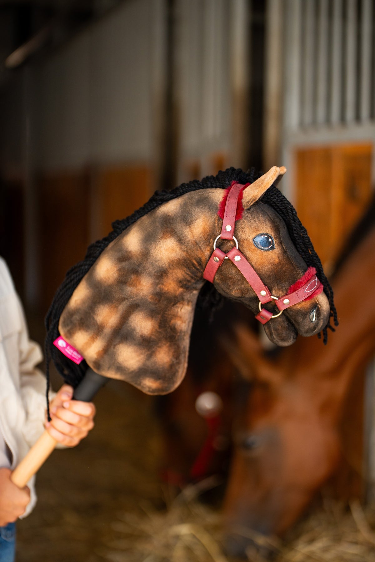Hlava Hobby Horse Stormy s černou ohlávkou od LarDen je zvednutá ve stáji, za ní je rozmazaný skutečný hnědák, s hnědým melírovaným plyšem, modrým okem a černou přízovou hřívou, s koženkovou kožešinovou ohlávkou Burgundy