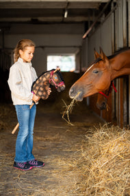 Lifestyle photo: a girl in a cream jacket holds the Hobby Horse Stormy with Black Halter by Hobby Horse LarDen up to a real bay horse eating hay in a stable - dappled brown plush head, black yarn mane - shown here with the Burgundy fur halter in leatherette.