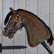 Hobby Horse Sundance s černou ohlávkou od Hobby Horse LarDen s plyšovým provedením, černou přízovou hřívou, uzdečkou a otěžemi, zachycující temperamentní vzhled anglického plnokrevného hřebce, upevněný na tyči na pozadí světlé stěny z dřevěných prken.