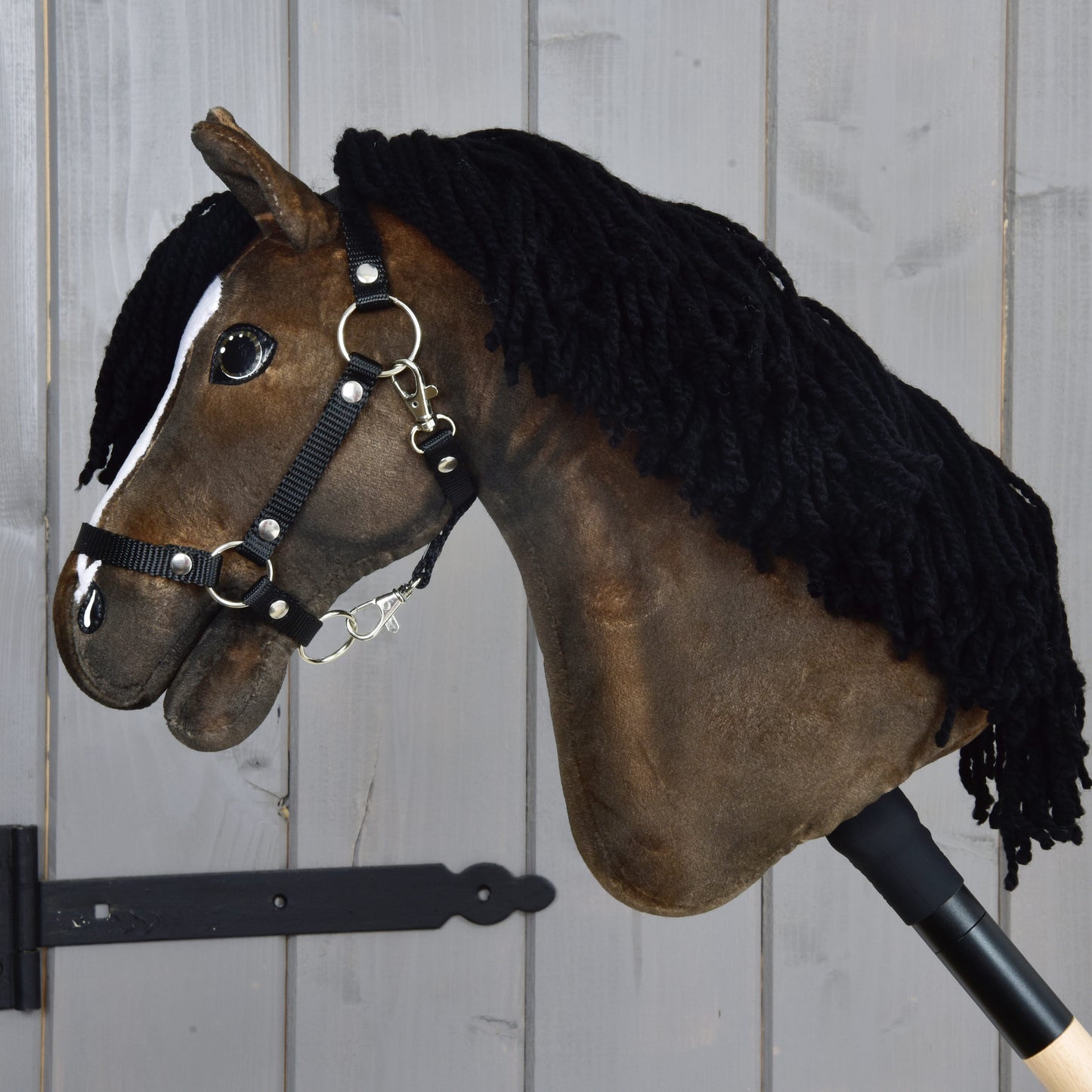 Hobby Horse Sundance s černou ohlávkou od Hobby Horse LarDen, hnědý látkový Hobby Horse inspirovaný rychlým plemenem anglický plnokrevník, s hustou černou přízovou hřívou, bílou lysinou na obličeji a černou nylonovou ohlávkou se stříbrnými přezkami. Je upevněný na tyči a zobrazený na šedém dřevěném pozadí.