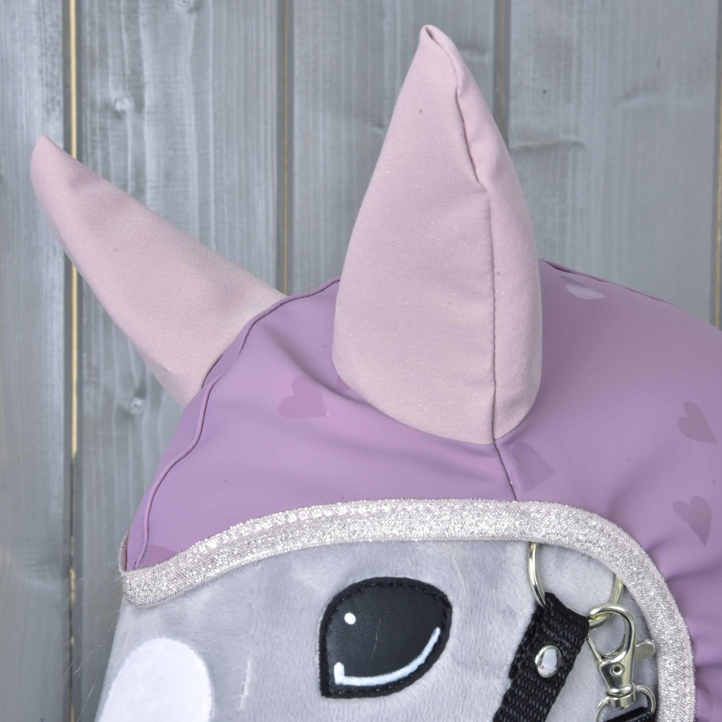 Detail hlavy plyšového koně Hobby Horse LarDen „Pláštěnka Sweetheart“ se světle fialovými ušima a potahem se srdíčkovým vzorem na dřevěném pozadí – půvabný doplněk pro Hobby Horse do každého počasí.