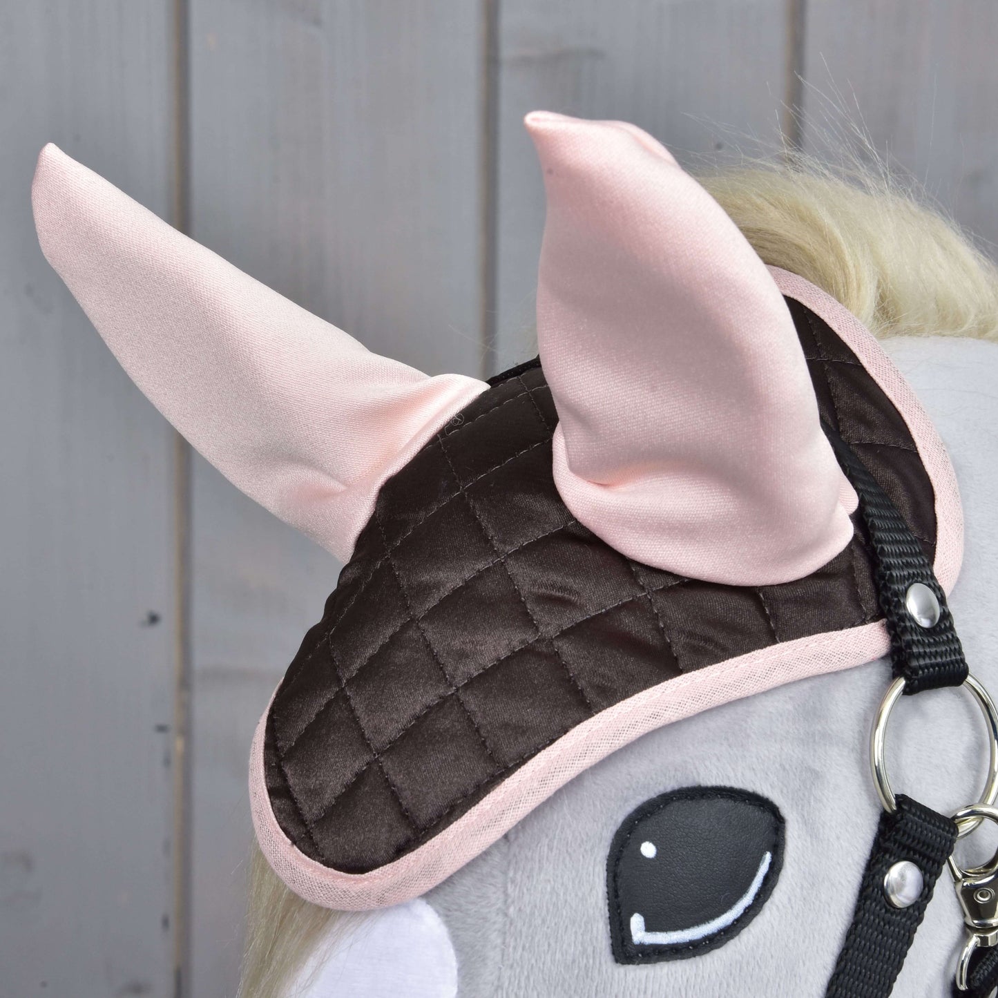 Detail Hobby Horse LarDen s čabrakou Chocolate, prošívanou hnědo-růžovou čabrakou se světle růžovými látkovými ušima pro ochranu proti hmyzu a s připevněnou uzdečkou, na pozadí dřevěného obložení.