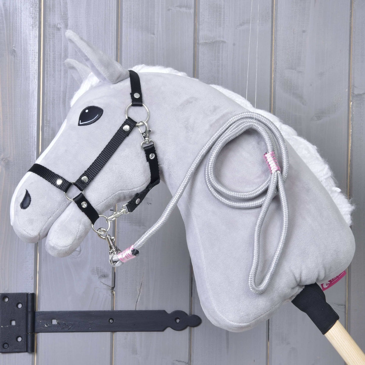Hobby Horse - vodítko Tizian od Hobby Horse LarDen v šedé barvě s černou uzdečkou, bílou hřívou a světlou dřevěnou tyčí opřené o šedou stěnu. Z krku visí stočené vodítko, ideální na hraní.