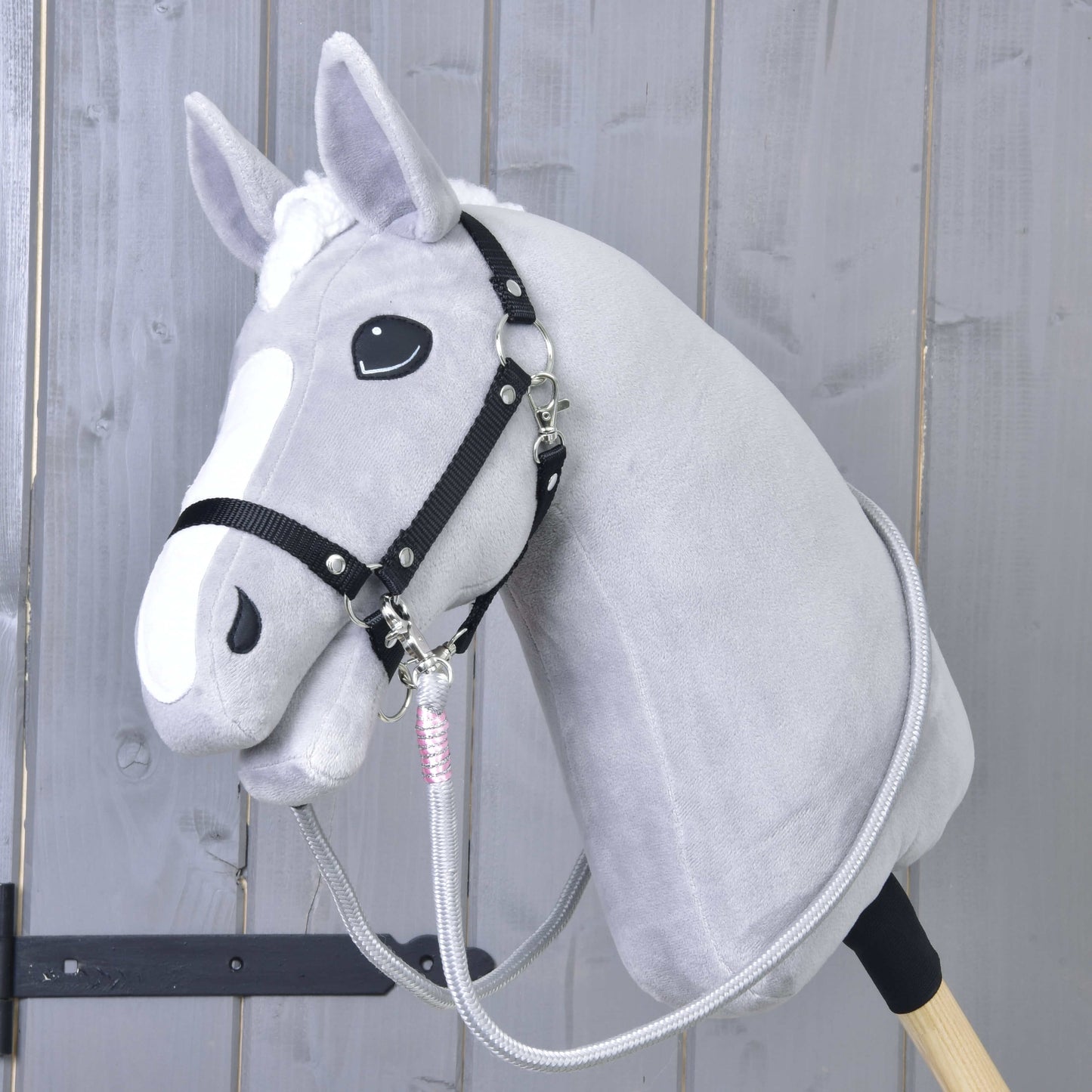 Hobby Horse LarDen Otěže Tizian na plyšovém šedém Hobby Horse s černou uzdečkou a ručně vyráběnými otěžemi, na dřevěné tyči před světlým dřevěným panelovým pozadím.