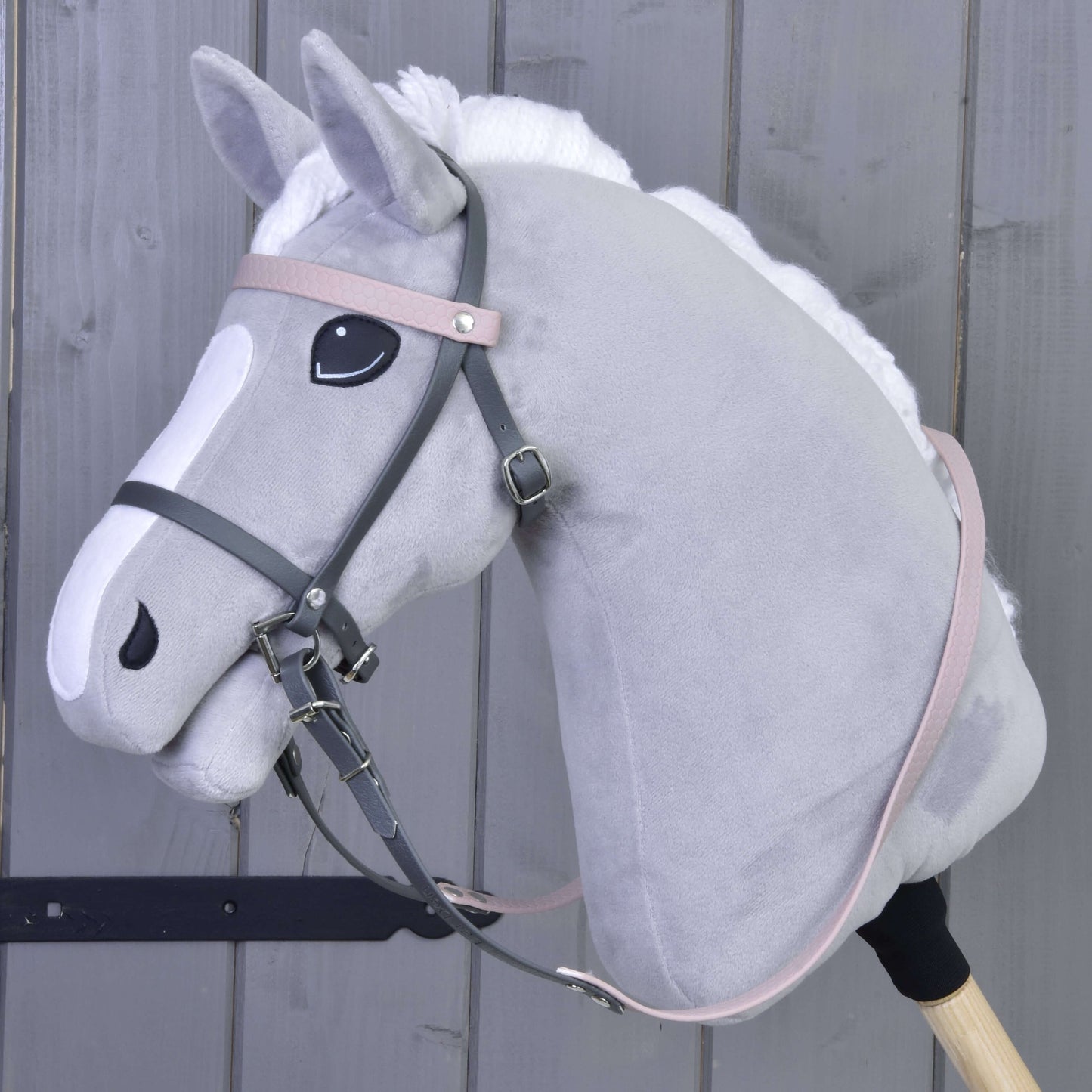 Plyšový šedý Hobby Horse s bílou hřívou a znaky na hlavě má Kompletní uzdečku šedou s růžovou čelenkou od Hobby Horse LarDen, je připevněný k dřevěné tyči a zobrazený na pozadí dřevěných panelů.