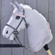Plyšový šedý Hobby Horse s bílou hřívou a znaky na hlavě má Kompletní uzdečku šedou s růžovou čelenkou od Hobby Horse LarDen, je připevněný k dřevěné tyči a zobrazený na pozadí dřevěných panelů.