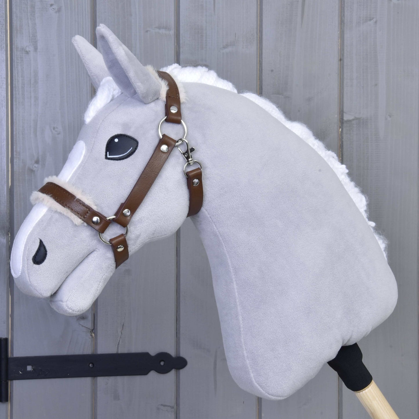 Ohlávka s beránkem z koženky Walnut od Hobby Horse LarDen na šedém plyšovém Hobby Horse s bílou hřívou, hnědou koženkovou uzdečkou a velkýma černýma očima na dřevěné tyči, opřeném o šedou dřevěnou stěnu.