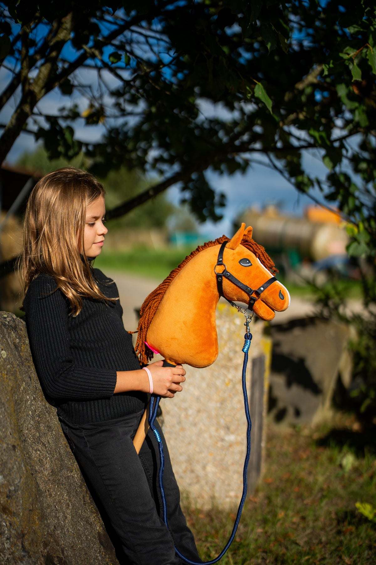 Dívka v černém topu se opírá o kamenný blok pod stromem a drží Hobby Horse Wendy s černou ohlávkou od LarDen, se světle ryzákovou plyšovou hlavou a zrzavou přízovou hřívou, s vodítkem Blues s pruhy z lana
