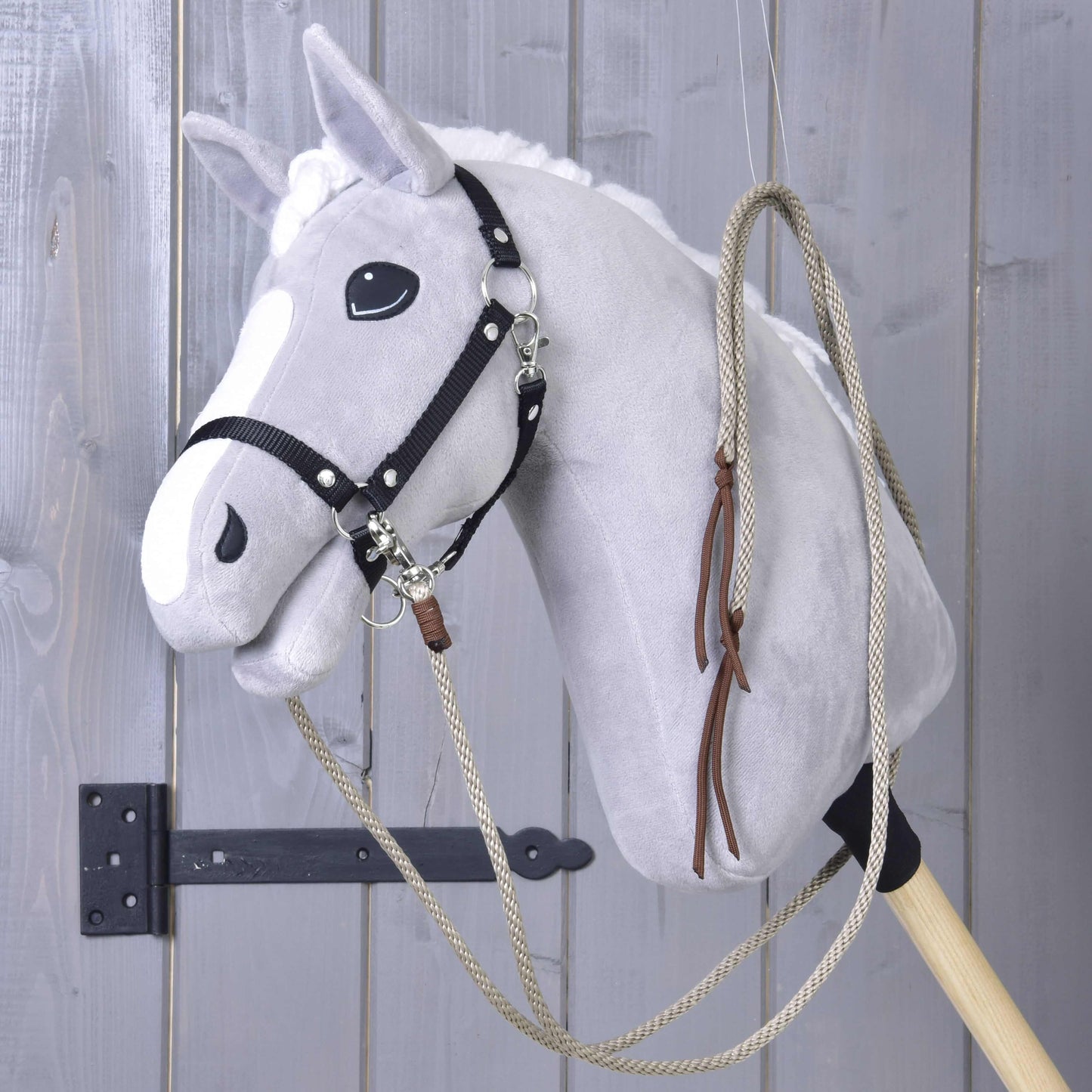 Šedý plyšový Hobby Horse od Hobby Horse LarDen s bílou hřívou, černým postrojem a Westernovými otěžemi Dallas rozdělenými, nasazený na dřevěné tyči a zavěšený před stěnou ze šedých dřevěných panelů.