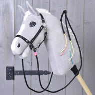 Hobby Horse LarDen "Westernové otěže Texas rozdělené", šedý plyšový koník s bílou hřívou, černou uzdečkou, dělenými westernovými otěžemi a barevnými stuhami na krku, nasazený na dřevěné tyči před šedými dřevěnými dveřmi.
