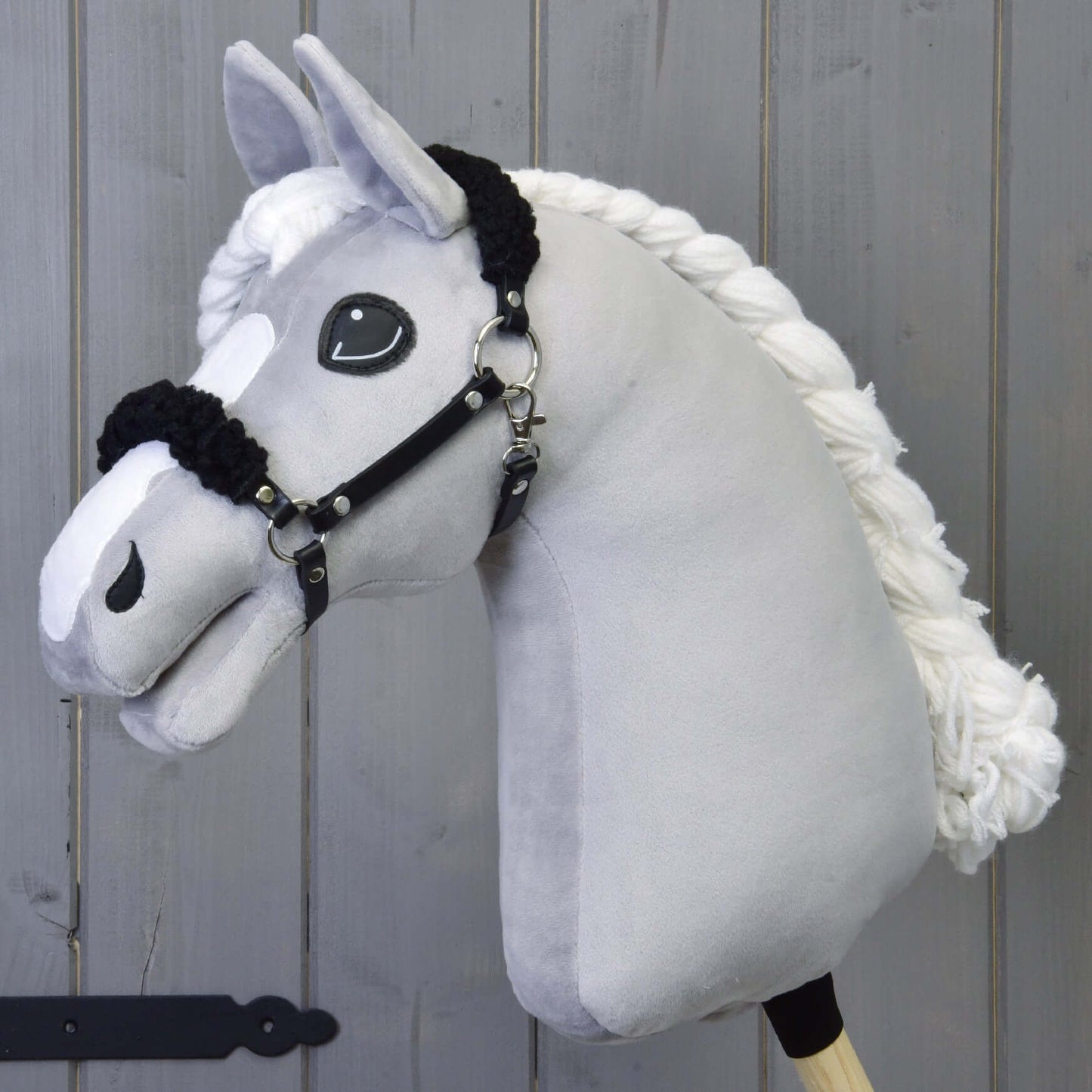 Černá ohlávka z koženky s černým beránkem od Hobby Horse LarDen na plyšovém světle šedém koni s černou koženkovou ohlávkou a velkýma vyšívanýma očima, před šedým dřevěným pozadím.