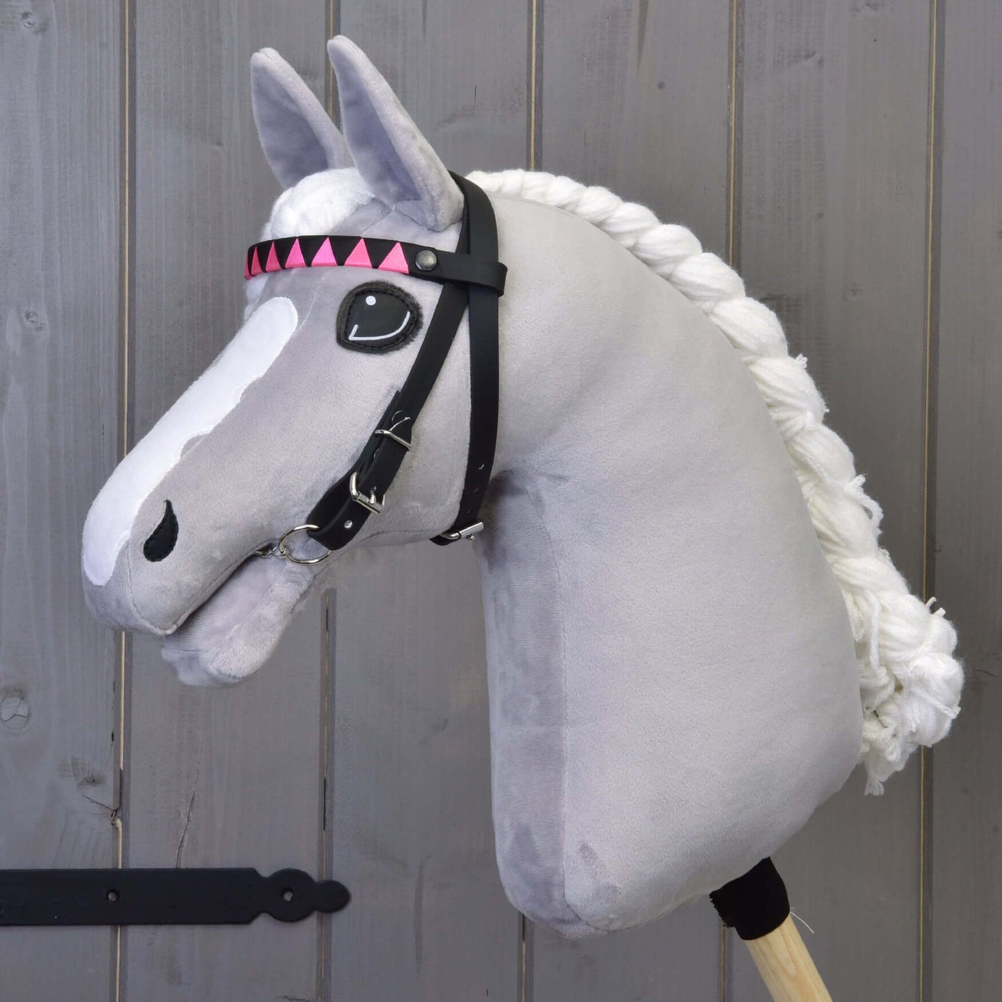 Uzdečka bez nánosníku růžová se 2 čelenkami (vel. M) od Hobby Horse LarDen s růžovými trojúhelníkovými prvky a odnímatelnými otěžemi, na šedém plyšovém Hobby Horse s bílou přízovou hřívou, před dřevěným pozadím.