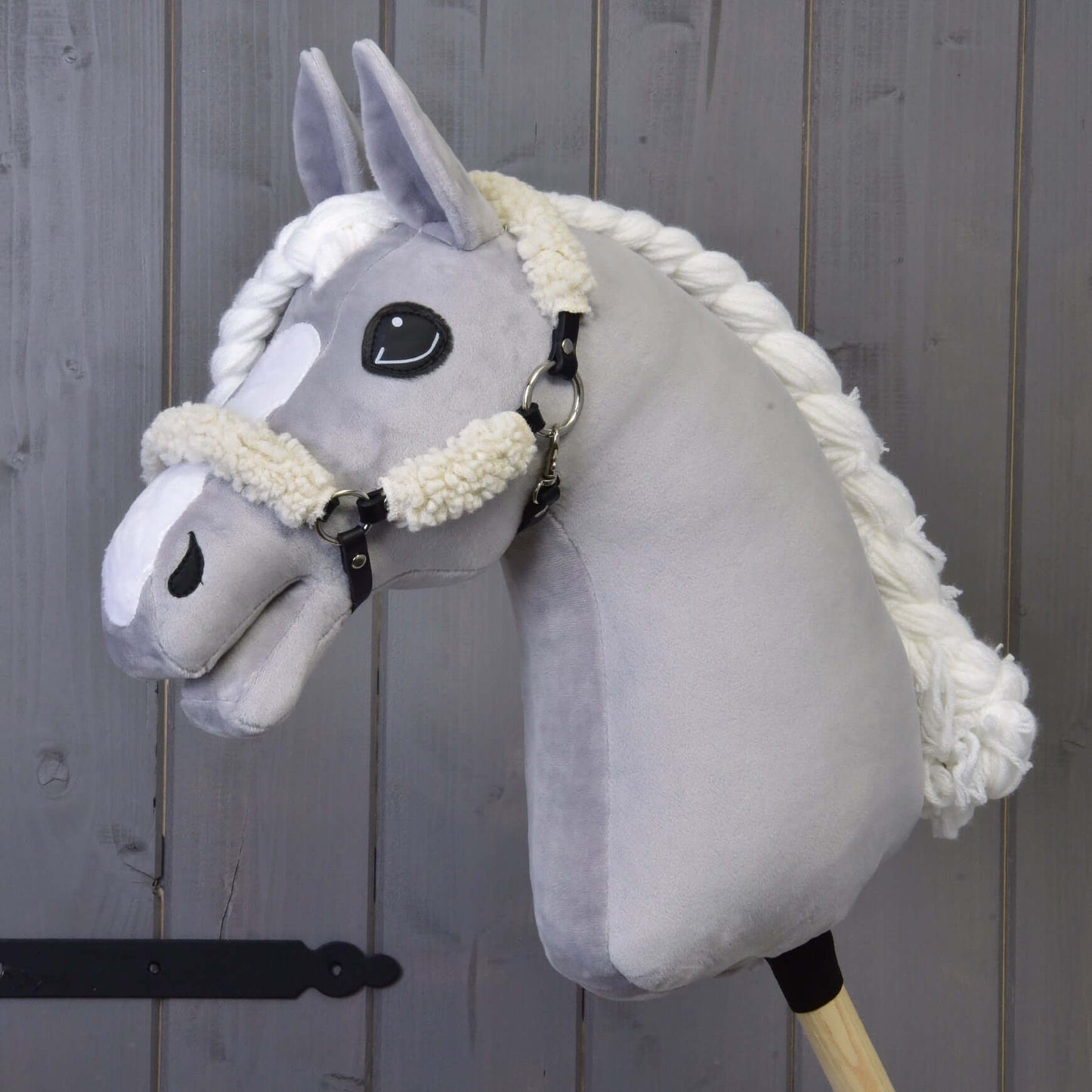 Plyšový šedý Hobby Horse od Hobby Horse LarDen s černou ohlávkou s kompletním beránkem a detaily z koženky, na dřevěné tyči před pozadím z dřevěných prken.