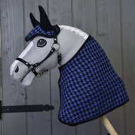 Hobby Horse LarDen s bílou hřívou a černou uzdečkou má na sobě Set slavnostní modrá, čabraku a deku se vzorem kohoutí stopy, a pózuje před šedou dřevěnou stěnou.