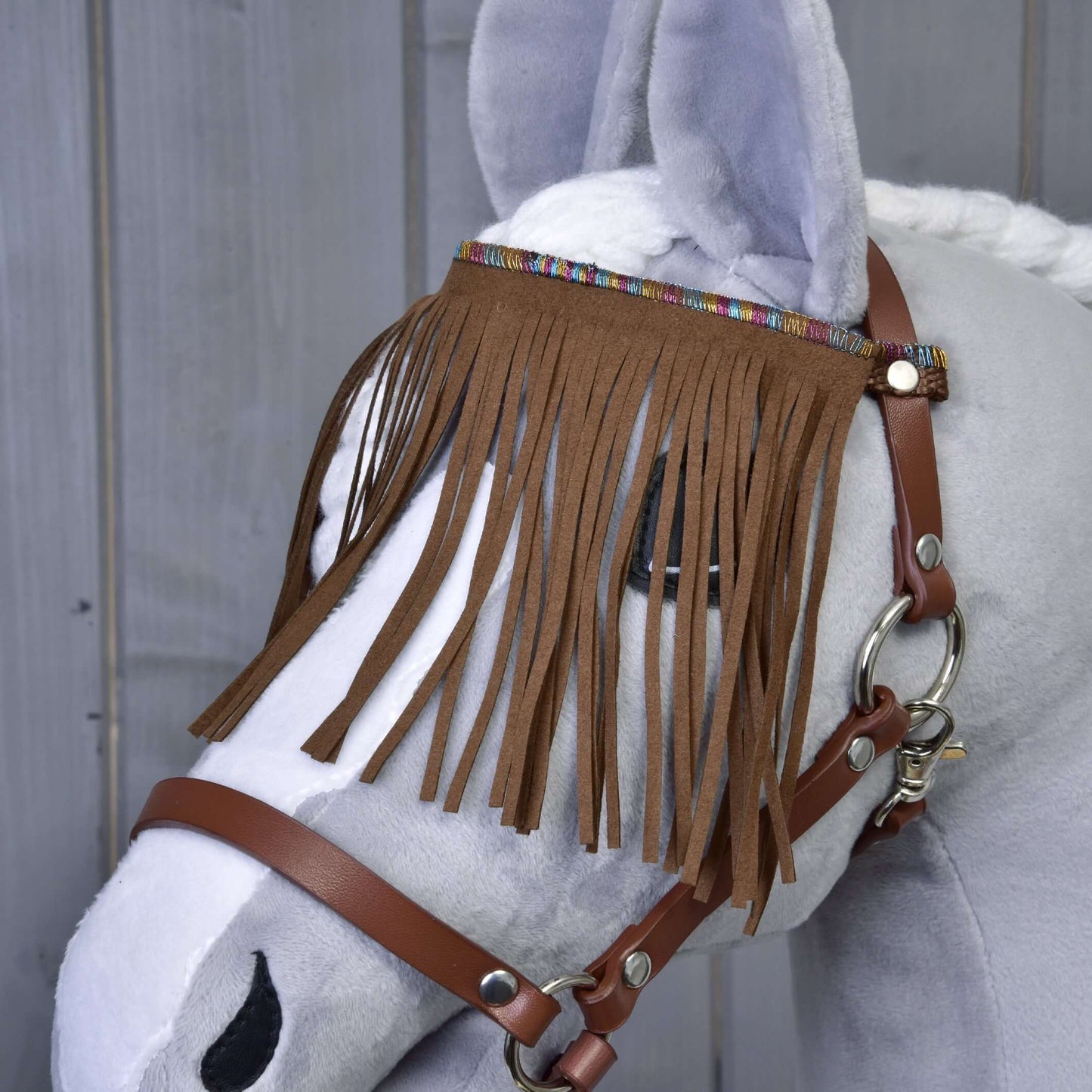 Ohlávka hnědá s třásněmi od Hobby Horse LarDen na šedém plyšovém Hobby Horse s bílou hřívou a ozdobnými třásněmi na čelence, na dřevěném pozadí.