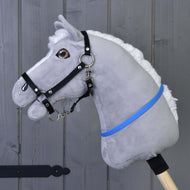 Hobby Horse nákrčník modrý (vel. S) od Hobby Horse LarDen na šedém plyšovém Hobby Horse s bílou hřívou, černou uzdečkou a modrofialovou stuhou na dřevěné tyči, před šedou dřevěnou stěnou.