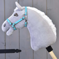 Šedý plyšový Hobby Horse s bílou hřívou a modrou uzdečkou má na sobě Ohlávku černou z koženky (vel. S) od Hobby Horse LarDen, na dřevěné tyči před šedými dřevěnými panely.
