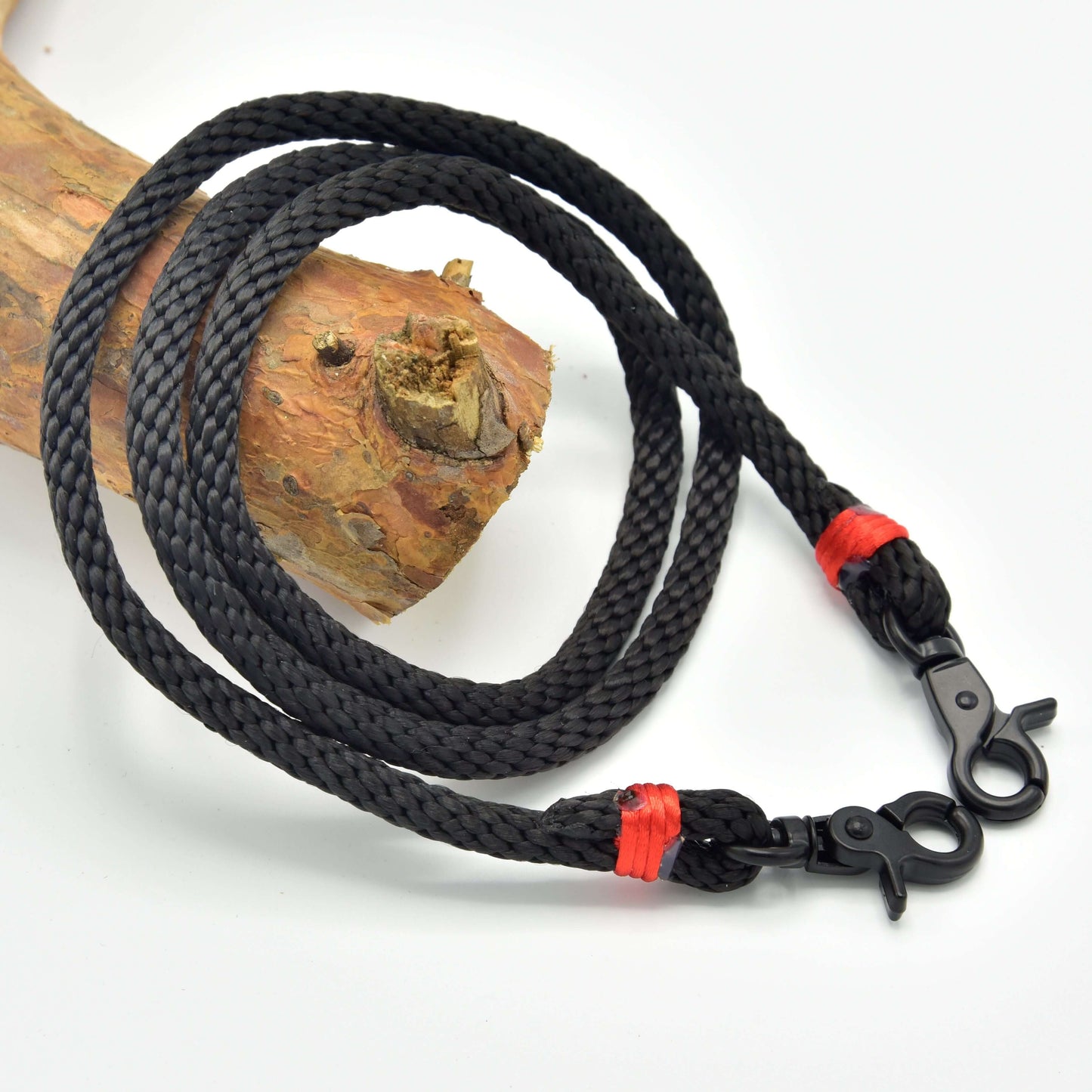 Otěže Jiskra od Hobby Horse LarDen z černého pleteného polypropylenového lana s kovovými klipy na obou koncích a červeným vázáním, stočené na dřevěné větvi; ideální jako ručně vyrobené otěže nebo otěže pro Hobby Horse.